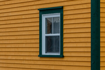 The exterior corner wall of a vibrant yellow colored wood clapboard siding. There's a single double hung window with green and white trim in the house. The corner of the building is thick green board.