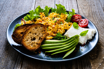 Breakfast - scrambled eggs with bacon, cottage cheese, avocado and vegetables on wooden table

