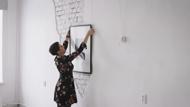 A woman gallerist hangs new paintings and collages on the wall of the exhibition hall. An expert on contemporary art works in galleries and prepares new exhibitions and auctions.