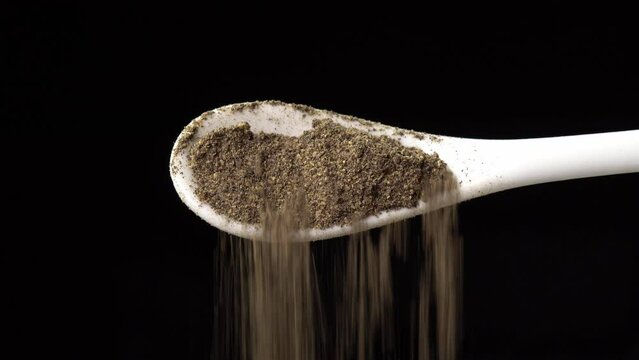 Ground black pepper on a spoon is poured out isolated on a black background. Dry seasoning in spoon. Spices and herbs for cooking, pepper powder