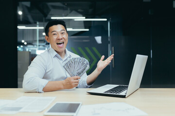 Young happy man businessman Asian holds cash in hands and rejoices. Sitting in the office at a table with a laptop, looking happily into the camera. He shows a winning gesture with his hand.