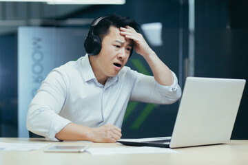 Upset and shocked young man in headphones sitting in office at desk over broken laptop. Holds his head, received bad news, problems with work.