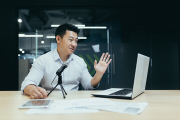 A young man sits in the office at the table, records a video, podcast, blog, webinar. Communicates online, gives interviews. Uses laptop and microphone.