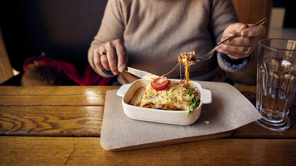 A woman eats lasagnia with cheese and a Behamel sauce from a white ceramic plate located next to a glass of water on the table