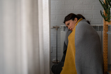 A woman in a cold room warming herself near a heater. Poor heating season. Cold adult woman covered...