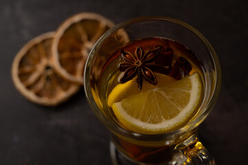 Close up of a glass teacup with black tea with lemon slices, star anise and cinnamon on a black metallic background.