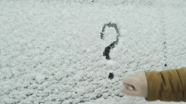A woman draws a question mark with her finger on white snow on a car window. Close-up.