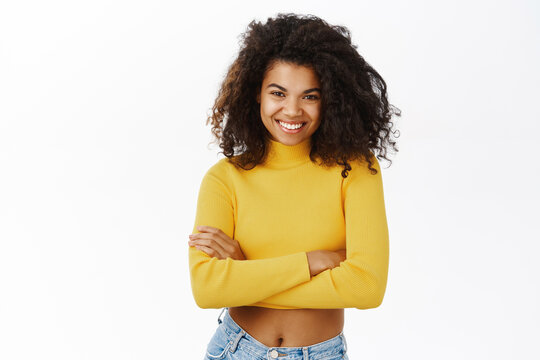 Beautiful young african woman, girl with curly hair, stylish outfit, laughing and smiling coquettish, posing in yellow crop top against white background