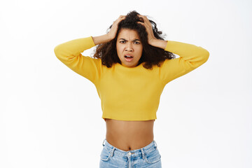 Portrait of shocked african american girl, looking troubled and frustrated, holding hands on head with scared face expression, white background