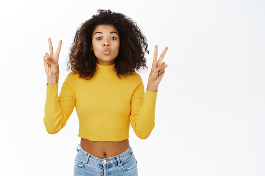 Positive african american girl shows peace sign, makes victory gesture, having fun, posing against white studio background