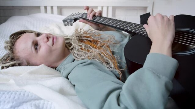 woman with dreadlocks on head singing song and playing acoustic guitar lying on bed in sunny cottage bedroom, simple living and hobby relaxing