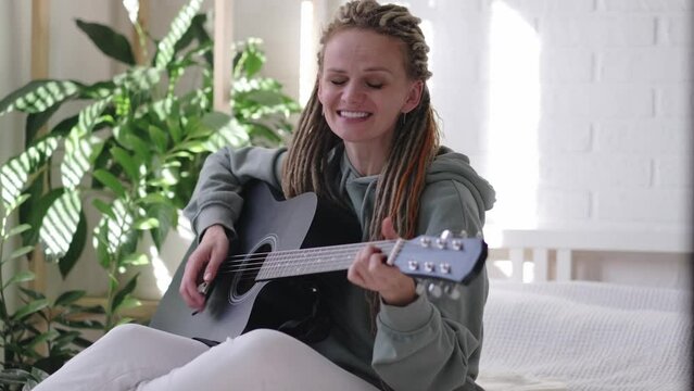 woman with dreadlocks on head singing song and playing acoustic guitar in sunny cottage bedroom, simple living and hobby relaxing