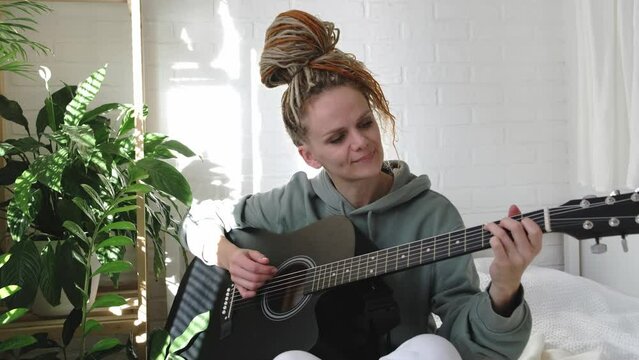 woman with dreadlocks on head singing song and playing acoustic guitar in sunny cottage bedroom, simple living and hobby relaxing