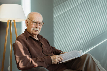 Senior man sitting alone on armchair in living room wearing glasses and holding book, looking thoughtfully at camera