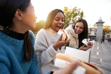 Group of cheerful Girls Eating Take Away Street Food while Sitting on the Bench in the in the beautiful Part of the City. Three Happy Girlfriends taking hot dogs in the Park
