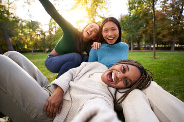 Naklejka premium Three multiracial girls lying on the grass taking a selfie looking at the camera. Group of cheerful friends having a fun together, sharing happiness on social media. Concept of friendship and free