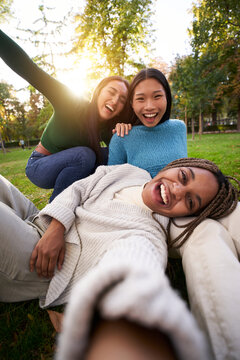 Vertical photo of Three multiracial girls lying on the grass taking a selfie looking at the camera. Group of cheerful friends having a fun together, sharing happiness on social media.