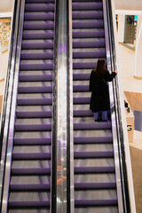 Faceless woman back view in a black coat standing on an escalator going up the stairs in a shopping mall, store, supermarket, or at the airport, train station. Modern stairway electronic system.