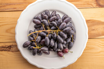 One bunch of juicy organic black grapes on a white ceramic platter on a wooden table, close-up, top view.