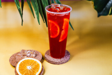 Red lemonade with ice and orange slices in a tall glass on a tropical background