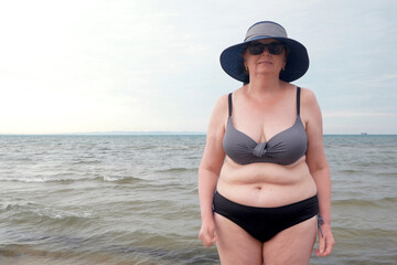 body positive senior woman in sunglasses wearing in swimsuit and summer haton the sea beach,