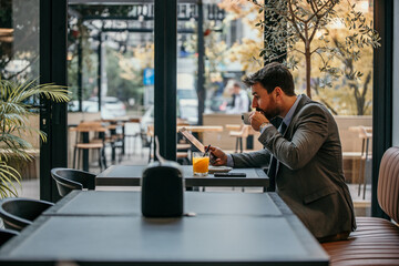 Side view of a businessman sitting alone next to the window, checking emails on the tablet and drinking a morning coffee.