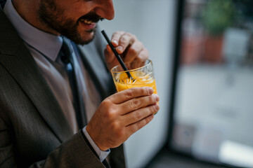 Close up image of a suited up businessman in a gray outfit standing in the office and drinking fresh orange juice on a straw.