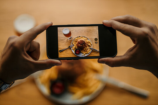 Top view of a male's hands taking a picture with a phone of his burger, french fries, and a beer pint. People photographing food concept. Focus on a phone