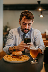 Focused businessman sitting in the local cafe, having a lunch break with a burger and beer and texting on his phone.