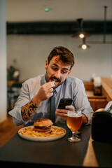Focused businessman sitting in the local cafe, having a lunch break with a burger and beer and texting on his phone.