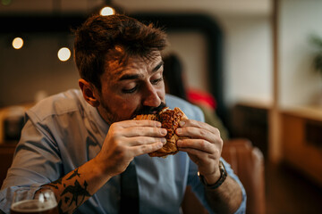 Bearded handsome businessman enjoying his lunch, biting a burger with a satisfied face.