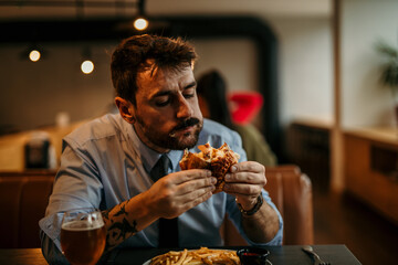 Bearded handsome businessman enjoying his lunch, biting a burger with a satisfied face.
