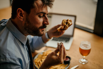 Close up image of a handsome bearded businessman wiped with a napkin eating his burger, drinking a beer, and working remotely on a laptop.