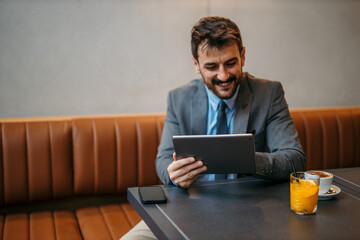 Good looking smiling businessman in a gray suit sitting in the cafe, using a digital tablet, drinking coffee and fresh orange juice. 