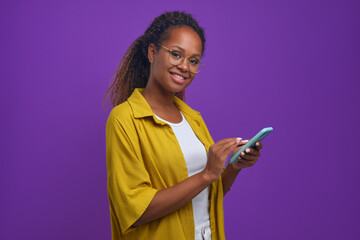 Young cheerful beautiful African American woman holding phone and looking at camera smiling runs fingers over display of smartphone making purchases via online application stands in lilac studio