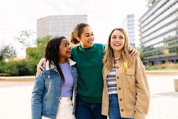 Three happy multiracial young women laughing outdoors. United female best friends having fun walking in city street. Community and international friendship concept