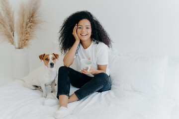 Happy attractive ethnic woman with curly hair wears white t shirt, jeans and socks, smiles pleasantly, drinks tea in comfortable bed, poses with dog, have lazy weekend. People, rest, animals concept