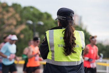 Rear view of policewoman with ponytail. Local police with reflective jacket with runners at the background.