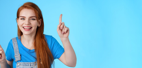 Fototapeta na wymiar Close-up cheerful carefree redhead dancing girl pointing up, raising fingers, smiling amused, having fun, stand blue background entertained, enjoy music party, disco mood, carefree emotions concept