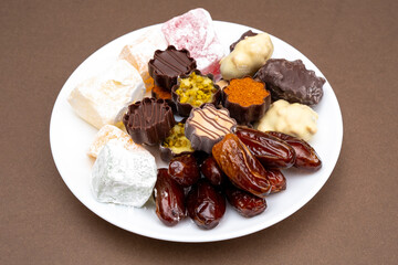 Valentine's Day White plate with sweets, dates, figs, chocolate, handmade nougat caramel. Festive brown background for Tu Bishvat Rosh Hashanah la'Ilanot.