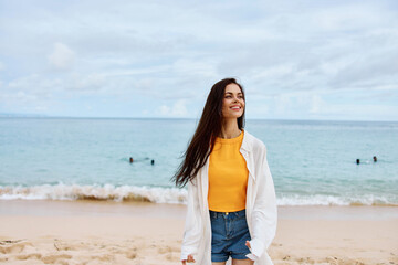 Fototapeta na wymiar Portrait of a happy woman smile with teeth with long hair brunette walks along the beach in a yellow tank top denim shorts and a white shirt by the sea summer travel and feeling of freedom, balance