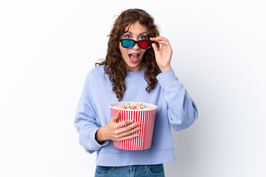 Young woman with curly hair isolated on white background surprised with 3d glasses and holding a big bucket of popcorns