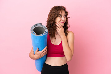 Young sport woman going to yoga classes while holding a mat isolated on pink background looking up while smiling