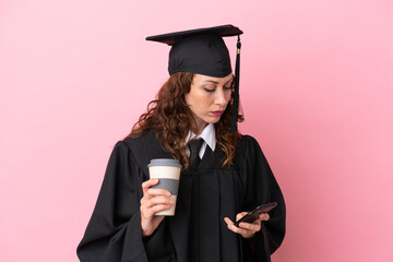 Young university graduate woman isolated on pink background holding coffee to take away and a mobile