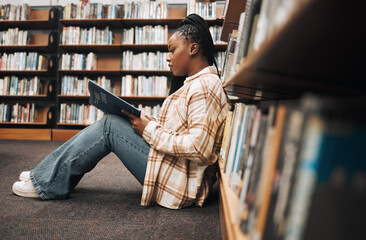 Library, college student and black woman reading books on ground floor for education, studying and...