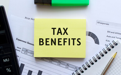 Text TAX BENEFITS on the page of a notepad lying on financial charts on the office desk.