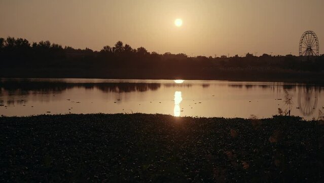 A beautiful sunset is reflected in the lake and with an observation wheel on the horizon and with vegetation around