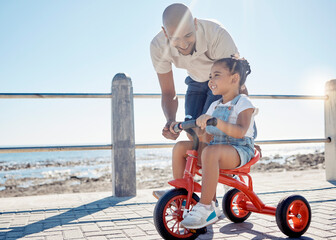 Kid, dad and learning to ride bicycle at beach promenade, sunshine and summer outdoors for fun, child development and play. Father teaching cycling to happy girl on bike at ocean sidewalk in nature