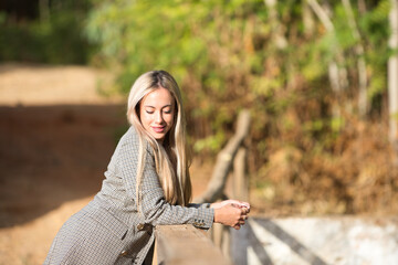 Obraz premium Beautiful blonde woman with straight hair is dressed in plaid jacket and denim skirt. The woman is happy and relaxed leaning on the railing of the wooden bridge in the forest and looks down.
