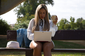 Portrait of Beautiful young blonde modern dressed woman sitting on a bench in the park with a laptop. The woman is holding an eco-friendly paper cup of coffee or infusion.